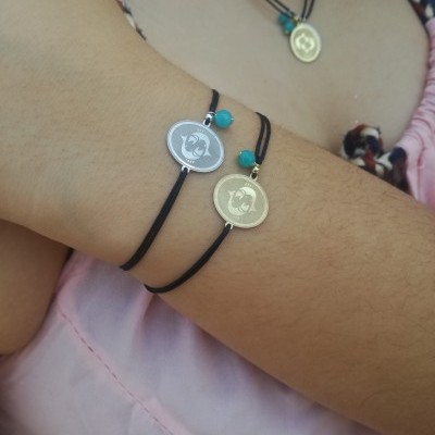 Pulseiras de fio preto com medalhas prata e dourada com contas azuis no pulso de uma pessoa com camisa rosa clara