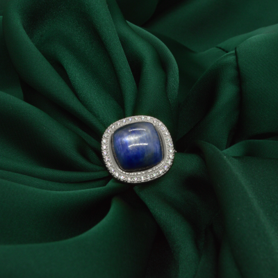 Anel com pedra azul quadrada em cabochão e cristais, sobre tecido verde escuro.
