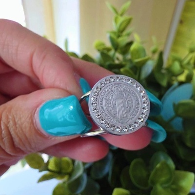 Anel prateado com medalha circular e strass segurado por mão com unhas azuis diante de plantas verdes