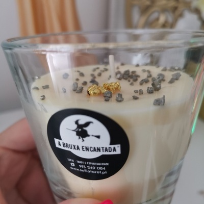 Vela branca em copo de vidro com pedras e detalhes dourados, etiqueta A BRUXA ENCANTADA.