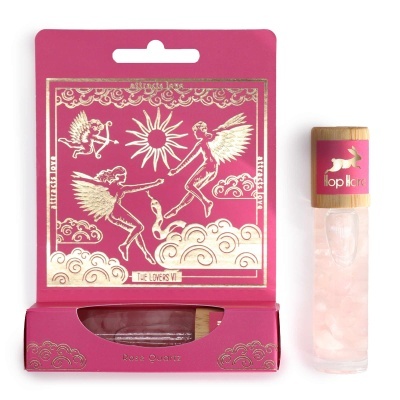 Perfume em frasco de vidro transparente com tampa de madeira, junto à embalagem decorada rosa e dourada com anjos e sol.