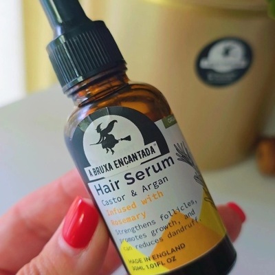 Frasco de sérum para cabelo com conta-gotas, rótulo com imagem de bruxa e texto, mãos segurando, planta em vaso ao fundo.