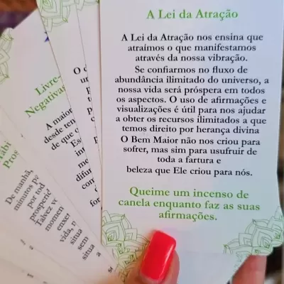 Mão segurando cartas com texto sobre Lei da Atração, bordas decorativas verdes, fundo de prateleiras desfocado