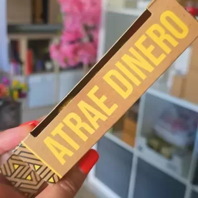 Embalagem dourada com texto ATRAE DINERO segurada por mão com unhas vermelhas
