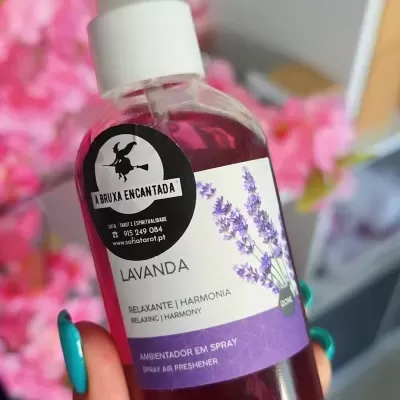 Frasco de ambientador spray lavanda com rótulo roxo e branco, em fundo com flores rosas desfocadas.