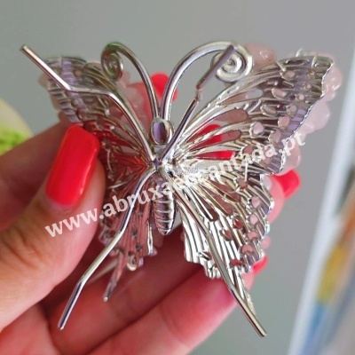Broche metálico em forma de borboleta prateada segurado por uma mão com unhas vermelhas