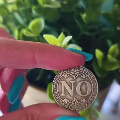 Moeda decorativa antiga com a palavra NO segurada por mão com unhas azuis e plantas ao fundo