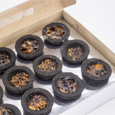 Tartes pequenas com base preta e recheio castanho com nozes dentro de caixa de cartão