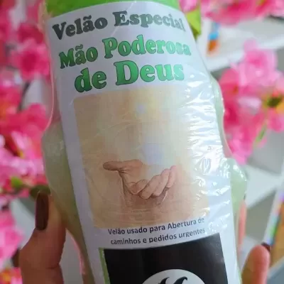 Vela verde com etiqueta branca e texto em verde, segurada por mão com unhas pintadas.
