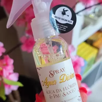 Frasco de spray Água Divina com rótulo branco e texto, decorado com flores cor-de-rosa ao fundo