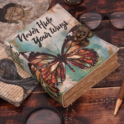 Caixa de livro decorativa com borboleta e texto 'Never Hide Your Wings' numa mesa de madeira com acessórios