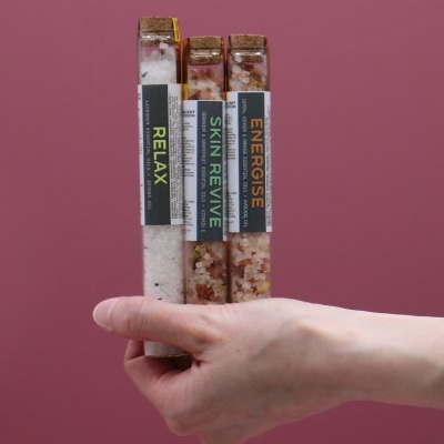 Três frascos de vidro com sais de banho e rótulos RELAX, SKIN REVIVE e ENERGISE numa mão