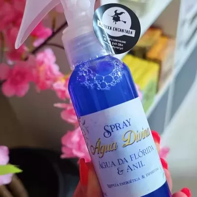 Frasco de spray azul com rótulo branco e texto em português, segurado por mão com unhas vermelhas, ao fundo flores rosa.
