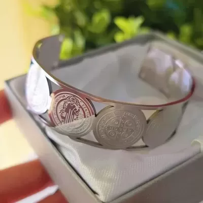 Pulseira metálica prateada com moedas religiosas dentro de caixa cinza acolchoada