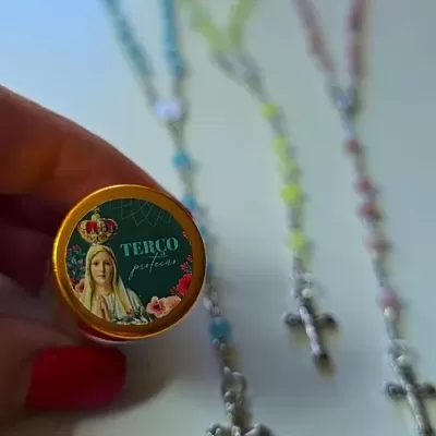 Frasco pequeno com tampa dourada e etiqueta 'TERÇO perfumado', terços coloridos com cruzes prateadas ao fundo