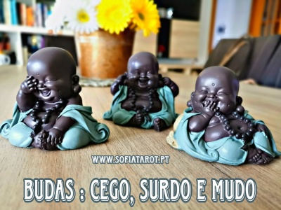 Três estatuetas de budas em madeira com roupas verdes, sobre mesa de madeira com vaso de flores ao fundo e texto promocional.