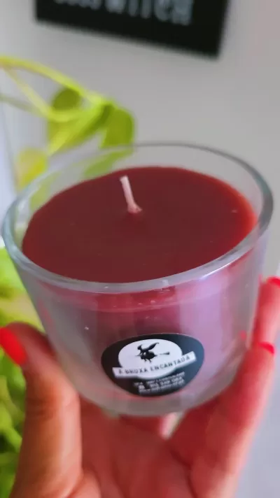 Vela vermelha em copo de vidro com etiqueta preta e planta ao fundo