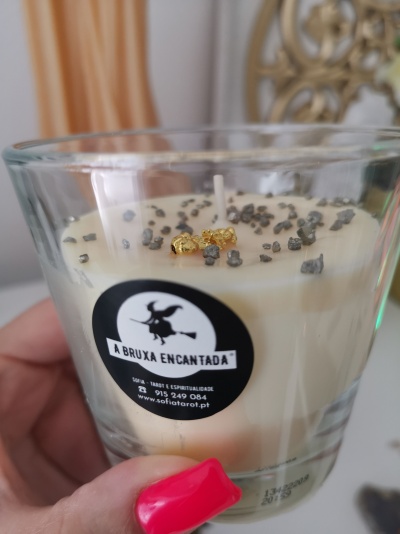 Vela branca em copo de vidro com pedras e detalhes dourados, etiqueta A BRUXA ENCANTADA.