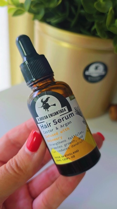 Frasco de sérum para cabelo com conta-gotas, rótulo com imagem de bruxa e texto, mãos segurando, planta em vaso ao fundo.