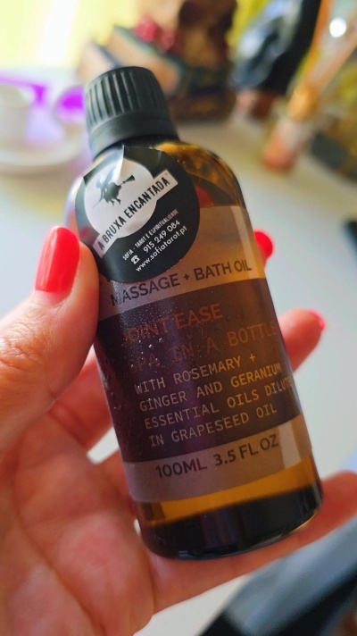 Frasco castanho de óleo para massagem com rótulo e tampa preta sendo segurado por uma mão com unhas pintadas de vermelho.