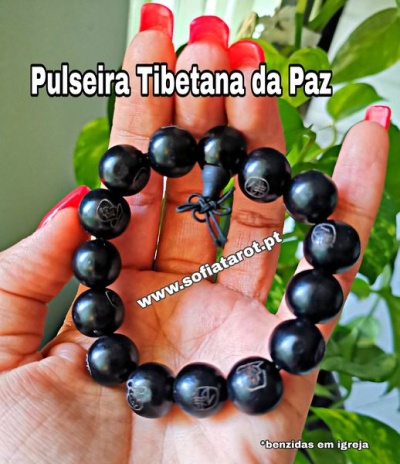 Pulseira Tibetana da Paz feita de contas pretas com símbolos, segurada por mão com unhas vermelhas, fundo de plantas