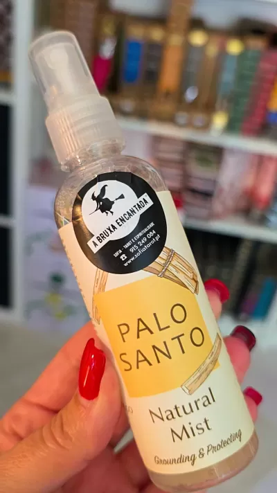Frasco de spray Palo Santo Natural Mist com rótulo amarelo e branco e adesivo preto, mão a segurá-lo com unhas vermelhas