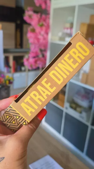 Embalagem dourada com texto ATRAE DINERO segurada por mão com unhas vermelhas