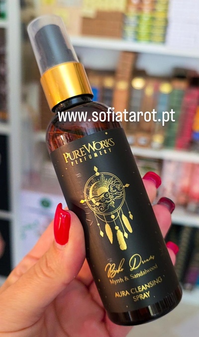 Frasco spray preto com rótulo dourado e tampa transparente numa mão com unhas vermelhas