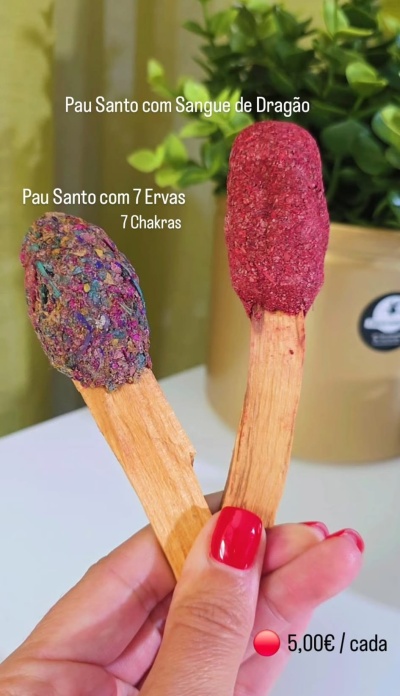 Dois pau santo com coberturas coloridas segurados por mão com unhas vermelhas, planta ao fundo.