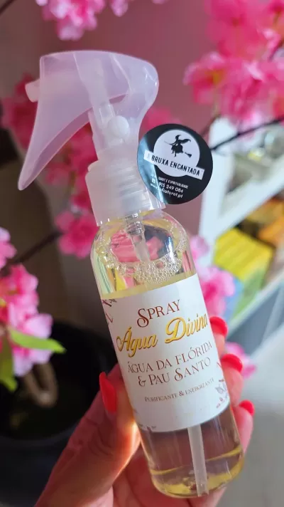Frasco de spray Água Divina com rótulo branco e texto, decorado com flores cor-de-rosa ao fundo