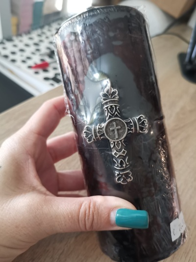 Vela preta decorativa com cruz metálica prateada, segurada por mão com unhas azuis sobre mesa