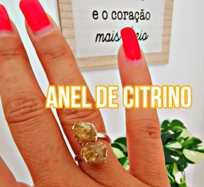 Anel de citrino com cristais amarelos em dedo com unhas vermelhas fluorescentes, fundo com planta e quadro com texto.