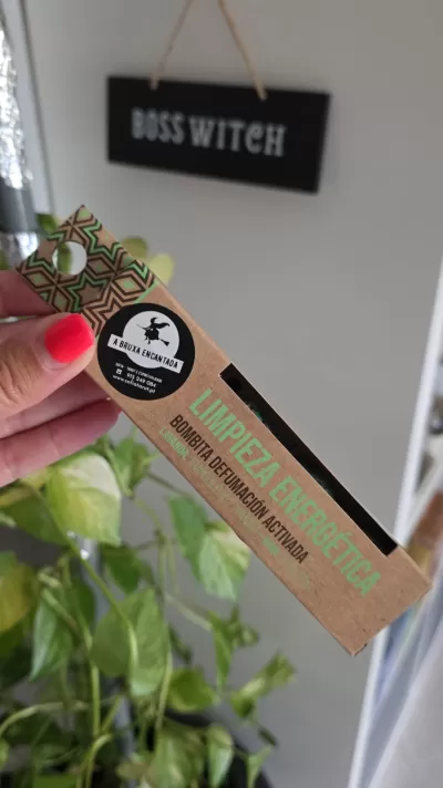 Embalagem de limpeza energética em papel kraft com texto e selo, segurada por mão com unhas vermelhas, fundo com placa BOSS WITCH e plantas