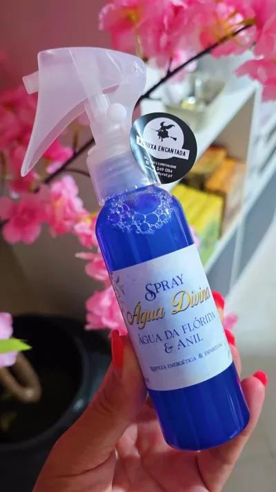 Frasco de spray azul com rótulo branco e texto em português, segurado por mão com unhas vermelhas, ao fundo flores rosa.