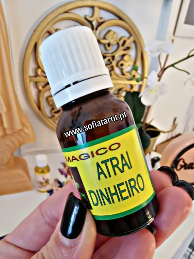 Frasco pequeno com rótulo amarelo que diz MAGICO ATRAI DINHEIRO segurado por mão com unhas pretas