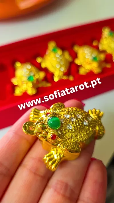 Figura de sapo dourado com pedras decorativas na mão, com caixa vermelha ao fundo e texto www.sofiat tarot.pt