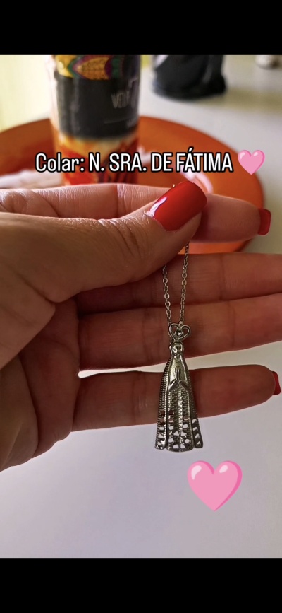 Colar filigranado de Nossa Senhora de Fátima em metal prateado segurado por mão com unhas vermelhas