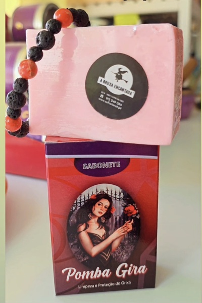Sabonete Pomba Gira em embalagem vermelha e roxa com imagem de mulher e barra de sabonete rosa com etiqueta preta da marca.
