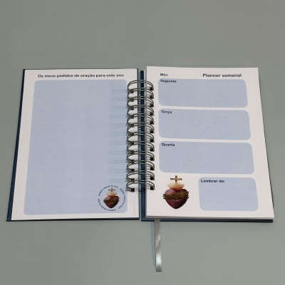 Planner semanal aberto com seções para dias da semana e pedidos de oração com ilustração de coração santo.