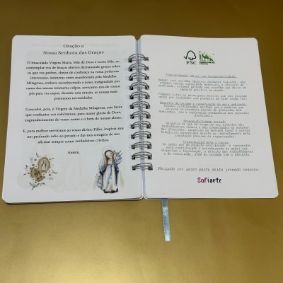 Livro de orações aberto com espiral, texto em português e selo FSC numa superfície amarela