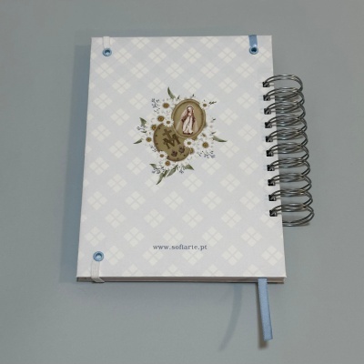 Caderno branco com padrão de losangos, ilustração de flores e figura religiosa, espiral metálica e marcador azul.