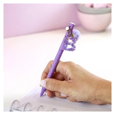 Caneta roxa com chaveiro letra B e pompom sobre papel quadriculado