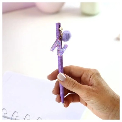 Caneta roxa com pompom lilás e letra N decorativa.