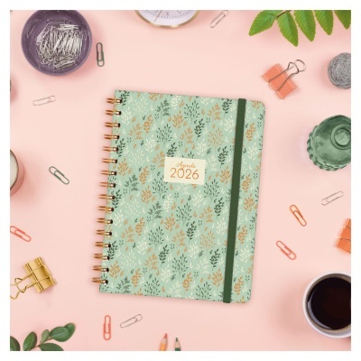 Agenda 2026 verde com padrão de folhas e elástico, rodeada por material de escritório sobre fundo rosa