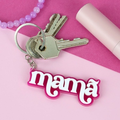 Chaveiro escrito mamã em branco com contorno rosa, junto a três chaves e objetos rosas e lilás no fundo.
