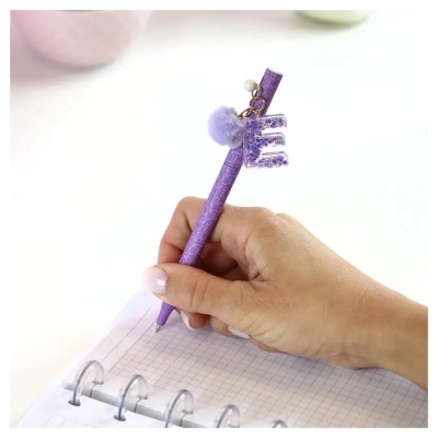 Caneta roxa com pendente letter E e pompom a escrever num caderno quadriculado.