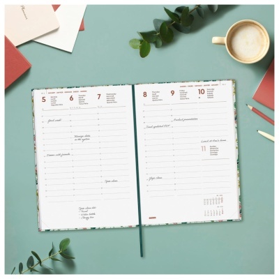 Agenda semanal aberta com capa floral e notas manuscritas, sobre mesa azul com caneta, lápis, café e plantas