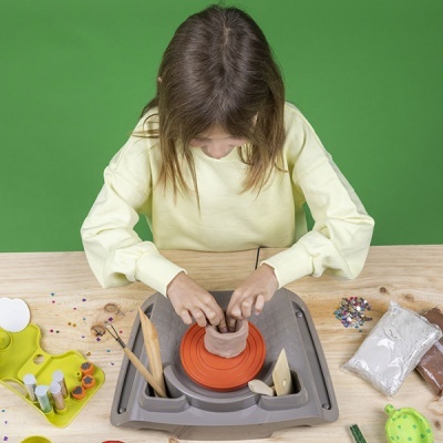 Criança a usar torno de olaria de plástico cinzento com disco vermelho para modelar barro castanho numa mesa com ferramentas e tintas.