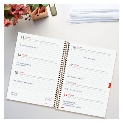 Agenda diária semanal aberta com texto em vermelho e azul, com capa espiral dourada sobre mesa branca