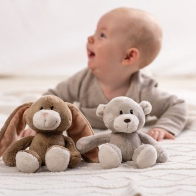 Dois peluches, um coelho e um ursinho, sobre uma manta bege com padrão geométrico, com um bebé ao fundo